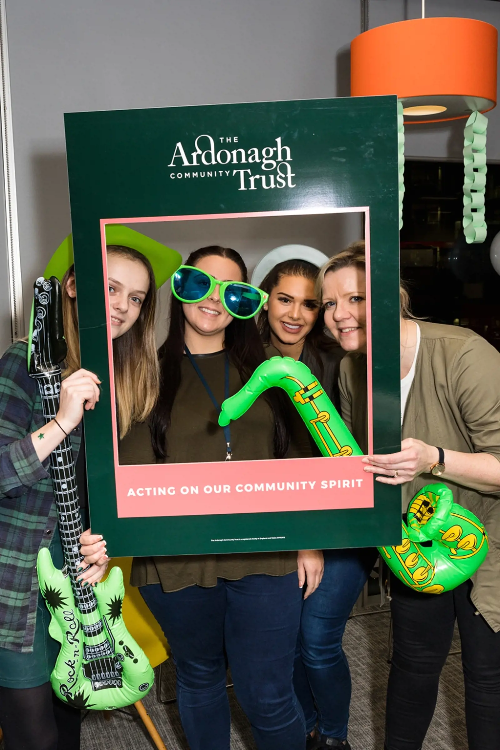 Four in photo booth style fancy dress, looking through Ardonagh's "Acting on our community spirit" cardboard frame